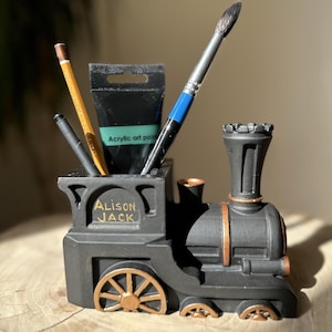 May include: Black toy train shaped desk organizer with gold accents. The train has a tube for holding pencils, paintbrushes, and a tube of acrylic paint. The train has the name "Alison Jack" written on the side.