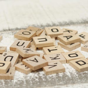 Könnte beinhalten: Eine Ansammlung von Holz-Scrabble-Steinen, die auf einer weißen, verwitterten Holzoberfläche verstreut sind. Die Steine zeigen schwarze Buchstaben und Zahlen, mit Buchstaben wie O, U, K, A und V. Das Bild ist sanft beleuchtet.