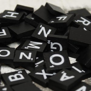 May include: A close-up shot of a pile of black Scrabble tiles. Each tile has a white letter and a point value. The tiles are scattered, creating a visually engaging arrangement. The letters are in various fonts.