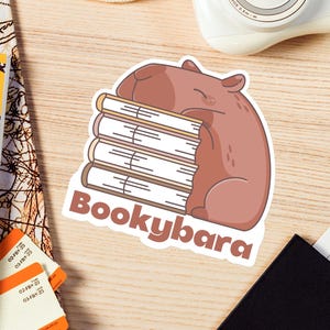 May include: A sticker featuring a brown capybara hugging a stack of books, with the word "Bookybara" below. The sticker is on a wooden surface, alongside a camera, travel documents, and a passport.