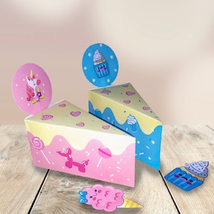 以下が含まれることがあります： 幻想的なデザインの、パステルカラーのケーキボックスが2つ。1つのボックスは、ユニコーン、ロリポップ、風船が描かれたピンク色。もう1つのボックスは、カップケーキとスプリンクルが描かれた青色。どちらのボックスも、白いフロスティングのデザインが施されています。