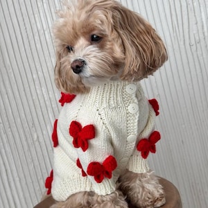 Puede incluir: Un perro pequeño con un cárdigan color crema con botones adornado con múltiples lazos rojos. El suéter tiene cuello y puños acanalados. El perro está sentado en un taburete de madera.