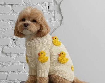 May include: A small dog wearing a cream-colored, button-down sweater adorned with yellow duck appliqués. The dog is sitting on a black stool against a white brick wall. The sweater has several yellow duck designs.