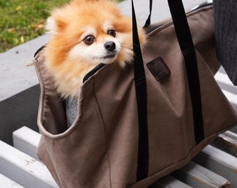 May include: A tan pet carrier with black straps holds a small, fluffy, orange and tan dog. The dog is looking to the side. The carrier has a small, square, brown label on the side.