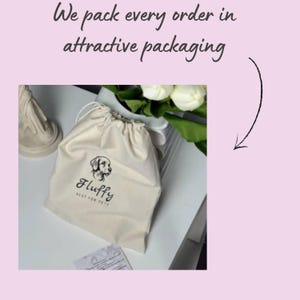 May include: A beige drawstring bag with a dog logo and the word "Fluffy" printed on it. The bag is on a white surface, with white flowers and greenery in the background. The text "We pack every order in attractive packaging" is at the top.