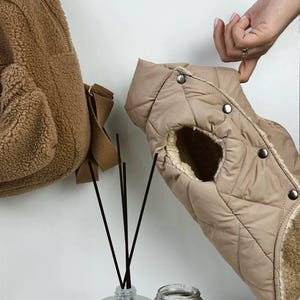 Puede incluir: Un chaleco acolchado beige para perros con forro de piel sint&eacute;tica se sostiene. Una mochila de oso de peluche marr&oacute;n cuelga de la pared. Un difusor y una vela est&aacute;n en una bandeja blanca.