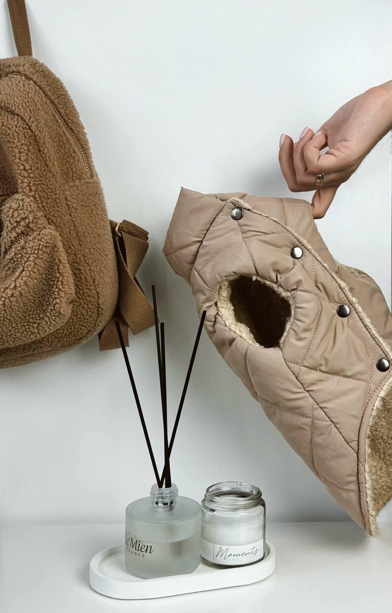 Puede incluir: Un chaleco acolchado beige para perros con forro de piel sint&eacute;tica se sostiene. Una mochila de oso de peluche marr&oacute;n cuelga de la pared. Un difusor y una vela est&aacute;n en una bandeja blanca.