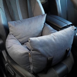 Puede incluir: Asiento de coche gris para mascotas, diseñado para su uso en un vehículo. El asiento está hecho de un material suave y afelpado y tiene una cremallera en la parte delantera y dos correas negras. El asiento proporciona comodidad y seguridad a las mascotas durante los viajes en coche.