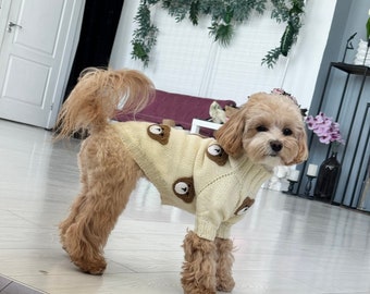 May include: A small, light brown dog wearing a cream-coloured sweater with brown bear faces. The sweater has a turtleneck and long sleeves. The dog is standing on a light brown wood floor.