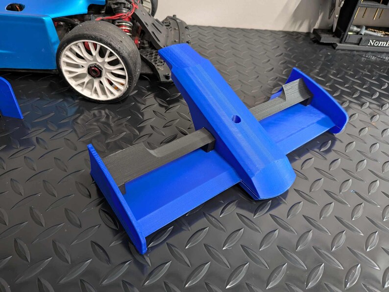 Pu&ograve; includere: Un'ala posteriore blu stampata in 3D per un'auto da corsa. L'ala ha due alette verticali e un'ala orizzontale con una barra di supporto nera.