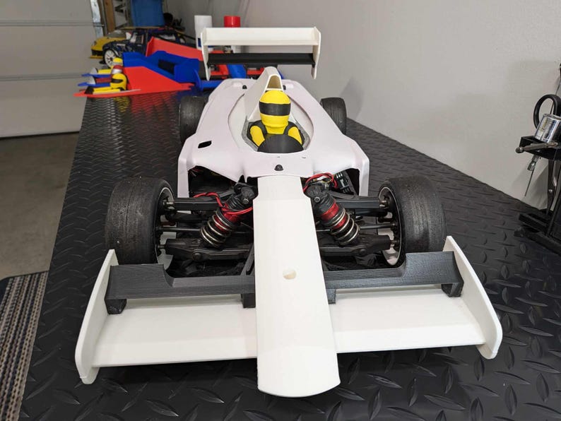 Pu&ograve; includere: Un'auto da corsa bianca con uno spoiler nero e una figura di pilota gialla. L'auto ha un'ala posteriore grande e un'ala anteriore.