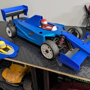 Pu&ograve; includere: Un'auto da corsa blu e bianca con un casco da pilota rosso. L'auto ha un grande alettone posteriore e un alettone anteriore pi&ugrave; piccolo. L'auto &egrave; seduta su una superficie nera e grigia.