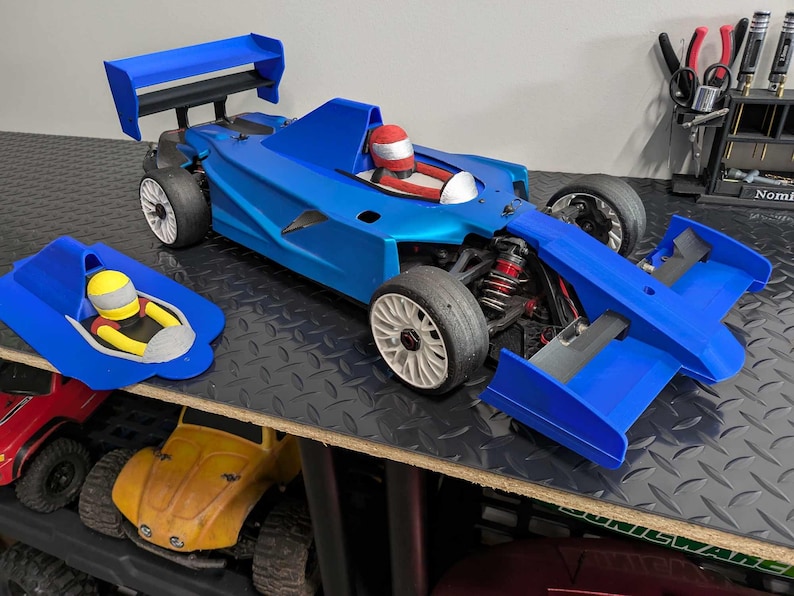 Pu&ograve; includere: Un'auto da corsa blu e bianca con un casco da pilota rosso. L'auto ha un grande alettone posteriore e un alettone anteriore pi&ugrave; piccolo. L'auto &egrave; seduta su una superficie nera e grigia.