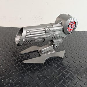 May include: A metallic gray, futuristic-looking device with a cylindrical end featuring red accents. The device has a complex, angular design with multiple components and is mounted on a stylized base. The object is likely a prop or model.