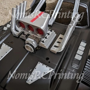 May include: A close-up of a black model car with a silver engine and exhaust pipes. The car has a detailed engine with a red stripe and silver accents. The car is sitting on a black surface.