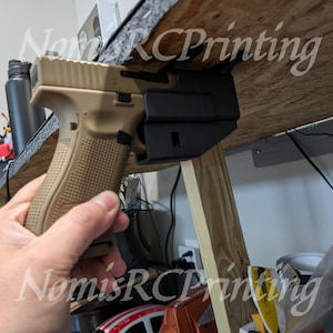 May include: A tan and black handgun is mounted to a wooden surface with a black plastic holster. The gun is held in place by a black plastic bracket. The text "Nomis RC Printing" is visible in the image.