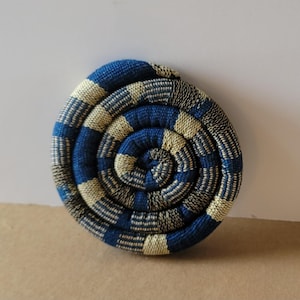 May include: A round, woven coaster with a spiral design. The coaster features a pattern of alternating blue and cream-colored squares. The coaster is made of a tightly woven material, and the overall design is symmetrical.