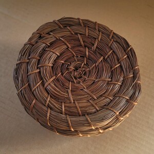K&ouml;nnte beinhalten: Ein runder, handgewebter Korb aus nat&uuml;rlichen braunen Fasern. Der Korb hat ein spiralf&ouml;rmiges Design mit aufwendiger Webart, wodurch eine strukturierte Oberfl&auml;che entsteht. Der Korb wird wahrscheinlich zur Aufbewahrung oder Dekoration verwendet.