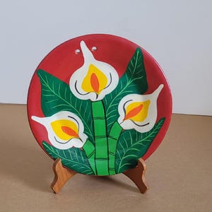 Könnte beinhalten: Ein handbemalter Keramikteller mit rotem Hintergrund und floralem Design. Das Design zeigt weiße Calla-Lilien mit gelben und orangefarbenen Zentren, grüne Blätter und einen grünen Stiel. Der Teller wird auf einem kleinen Holzständer präsentiert.