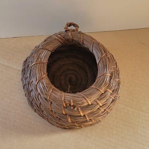 K&ouml;nnte beinhalten: Ein handgewebter, runder Korb mit einer kreisf&ouml;rmigen &Ouml;ffnung. Der Korb besteht aus braunen Naturfasern und hat oben eine kleine Schlaufe zum Aufh&auml;ngen. Das Gewebe ist eng, was ihm ein robustes, rustikales Aussehen verleiht. Der Korb ist leer.