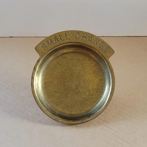 Könnte beinhalten: Ein Vintage-Münzschale aus Messing mit den Worten "SMALL CHANGE" auf der Oberseite geprägt. Die Schale ist rund mit einem leicht erhöhten Rand und einem kleinen, gebogenen Griff. Das Messing hat eine warme, gealterte Patina.