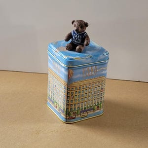 May include: A blue and yellow tin box with a picture of a building and hot air balloons. A small brown teddy bear wearing a blue shirt with the text "Saks Fifth Avenue" is sitting on top of the box.