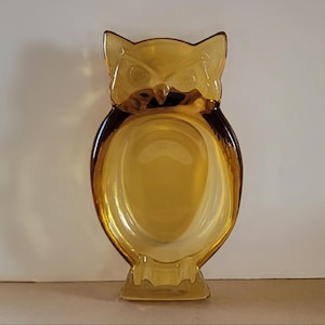 Könnte beinhalten: Bernsteinfarbene Glasschale in Eulenform. Die Schale hat einen breiten, ovalen Körper mit einer flachen Schüssel und einem detaillierten Eulengesicht oben. Das Glas hat eine glänzende Oberfläche und ist wahrscheinlich Vintage.