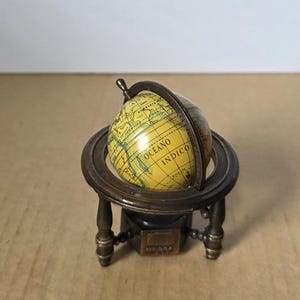 Puede incluir: Un pequeño globo terráqueo esférico de color amarillo con detalles geográficos impresos y el texto Oceano Indico, montado en un marco circular metálico oscuro sostenido por cuatro patas decorativas y una base central con placa.