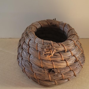 K&ouml;nnte beinhalten: Ein handgewebter, brauner Korb mit runder Form und offener Oberseite. Der Korb besteht aus d&uuml;nnen, naturfarbenen Fasern, die eng miteinander verwoben sind. Das Design weist ein dekoratives Knotendetail auf der Vorderseite auf.