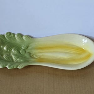 May include: A ceramic serving dish shaped like a head of endive or bok choy. The vegetable-shaped platter features textured, light green leaves at one end that transition into a smooth, elongated, pale yellow and cream-colored concave center.