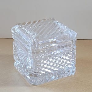 Könnte beinhalten: Eine transparente, quadratische Glas-Schmuckschatulle mit Deckel. Die Box hat ein sich wiederholendes diagonales Muster auf allen Seiten, das einen strukturierten Effekt erzeugt. Der Deckel spiegelt das Muster wider. Die Box ist für kleine Gegenstände konzipiert.