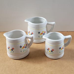 May include: Three white ceramic jugs with a floral design. The jugs are decorated with small yellow, orange, and blue flowers. The jugs are different sizes.