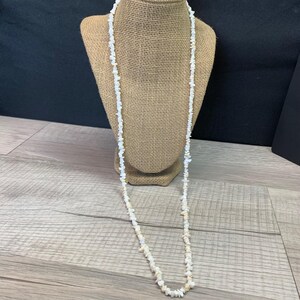 Peut inclure: Un long collier à brin unique composé de petites pierres concassées irrégulières de couleur blanche et blanc cassé. Le collier présente un design continu et uniforme sans fermoir visible, affichant un aspect naturel et texturé sur toute la longueur.