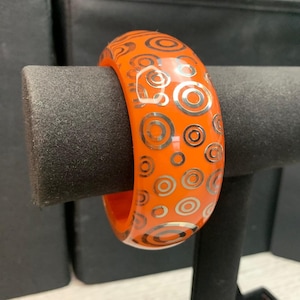 May include: A bright orange bangle bracelet featuring a repeating pattern of concentric silver circles of varying sizes. The bracelet has a smooth, glossy finish and a thick, rounded profile, showcasing a bold contrast between the orange and silver.