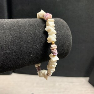 May include: A beaded bracelet featuring irregular, flat, off-white shell chips interspersed with small, smooth, light purple shell fragments and tiny, round, metallic gold-coloured spacer beads arranged in a repeating pattern along the strand.
