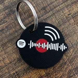 Op de afbeelding: Zwarte ronde sleutelhanger met een Spotify-code ontwerp. De sleutelhanger heeft een wit Spotify-logo, witte geluidsgolven en een wit audio golfvorm ontwerp met een rode kern. Een zilveren metalen ring is bevestigd.