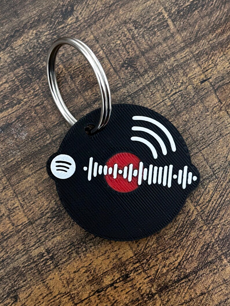 Op de afbeelding: Zwarte ronde sleutelhanger met een Spotify-code ontwerp. De sleutelhanger heeft een wit Spotify-logo, witte geluidsgolven en een wit audio golfvorm ontwerp met een rode kern. Een zilveren metalen ring is bevestigd.