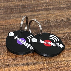 Op de afbeelding: Twee zwarte vinyl sleutelhangers met zilveren ringen. De ene heeft een paars midden, de andere een rood midden. Beide hebben witte geluidsgolfontwerpen. Op een houten oppervlak.