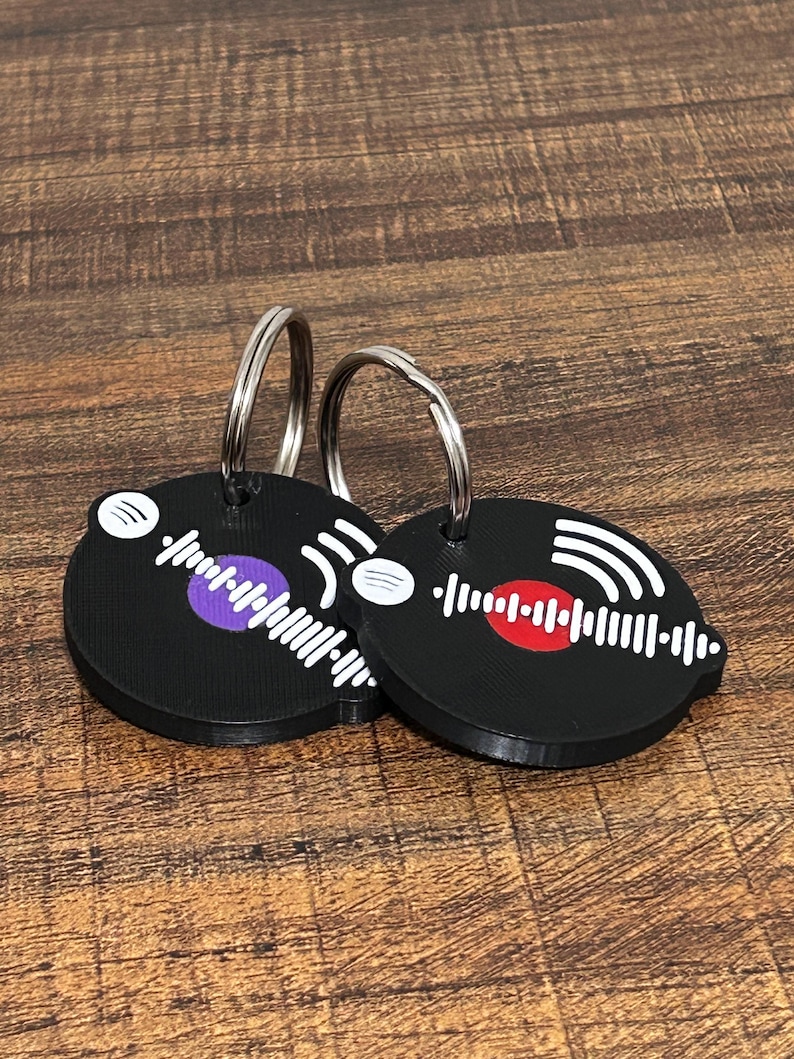 Op de afbeelding: Twee zwarte vinyl sleutelhangers met zilveren ringen. De ene heeft een paars midden, de andere een rood midden. Beide hebben witte geluidsgolfontwerpen. Op een houten oppervlak.