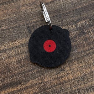 Op de afbeelding: Zwarte vinylplaat sleutelhanger met een rode middenlabel en een zilveren sleutelring. De sleutelhanger is rond en heeft een gestructureerd oppervlak, dat lijkt op een miniatuurplaat. Een leuk accessoire voor muziekliefhebbers.
