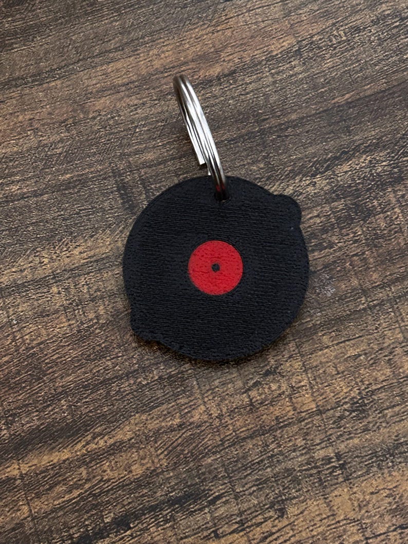Op de afbeelding: Zwarte vinylplaat sleutelhanger met een rode middenlabel en een zilveren sleutelring. De sleutelhanger is rond en heeft een gestructureerd oppervlak, dat lijkt op een miniatuurplaat. Een leuk accessoire voor muziekliefhebbers.