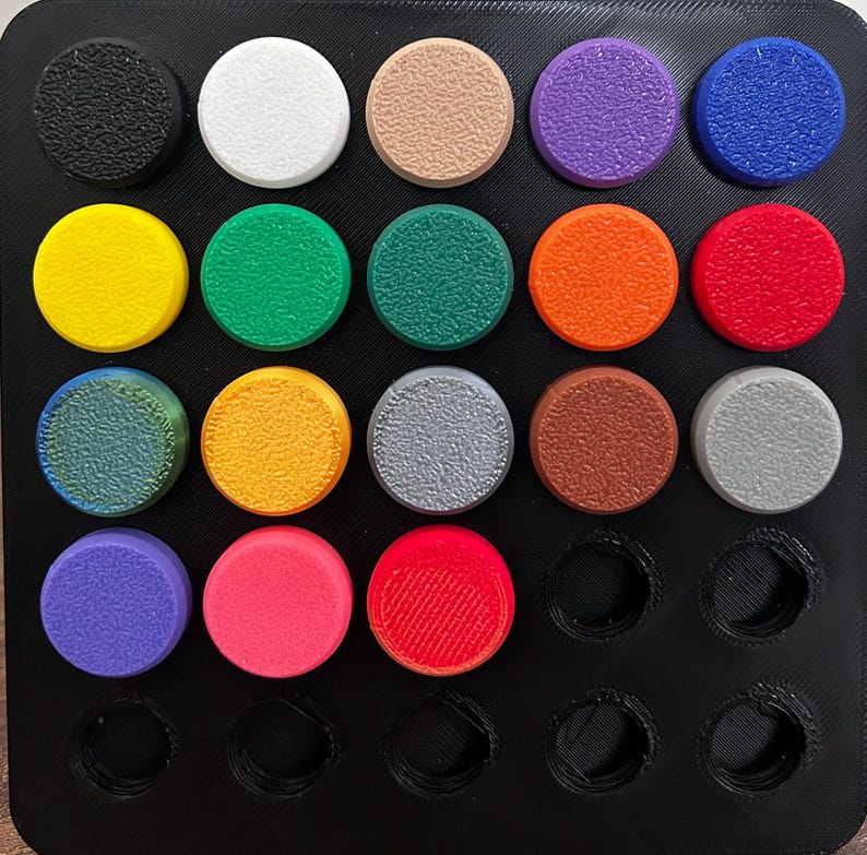 Op de afbeelding: Een zwarte tray met 18 ronde, getextureerde objecten in verschillende kleuren. De kleuren zijn onder meer zwart, wit, beige, paars, blauw, geel, groen, oranje en rood. De tray heeft drie lege ronde ruimtes.