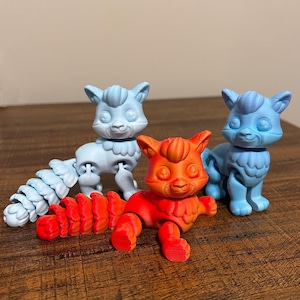 May include: Three articulated cat figurines in shades of blue and orange are displayed on a wooden surface. The cat toys have detailed features and poseable joints. One cat is orange, while the other two are blue.