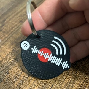 Op de afbeelding: Zwarte sleutelhanger met een cirkelvormig ontwerp met een rode kern, witte geluidsgolf en Spotify-logo. De sleutelhanger is bevestigd aan een zilveren metalen ring.