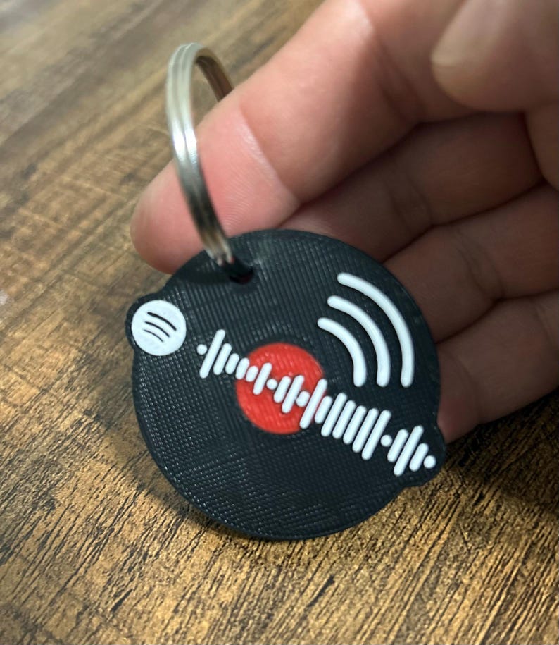 Op de afbeelding: Zwarte sleutelhanger met een cirkelvormig ontwerp met een rode kern, witte geluidsgolf en Spotify-logo. De sleutelhanger is bevestigd aan een zilveren metalen ring.