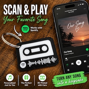 May include: A black and white rectangular keychain shaped like a cassette tape with a Spotify code, attached to a silver metal ring. Next to it, a smartphone displays a music player interface with the song title Our Song and a sunset beach image.
