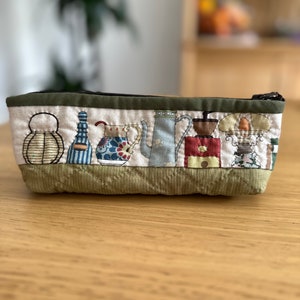 May include: A green quilted zippered pouch with a white fabric panel featuring a row of colourful illustrations of teapots, coffee pots, and other kitchen items.