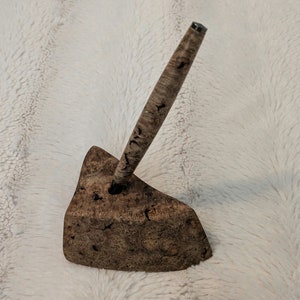 May include: A wooden pen holder with a unique, natural wood grain pattern. The holder is shaped like a triangle and has a small hole in the top for holding a pen. The pen is made of wood and has a silver top.