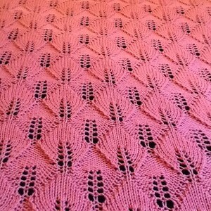 May include: A pink knitted blanket with a lacy, leaf-like pattern. The blanket is made with a soft yarn and has a delicate, feminine feel.