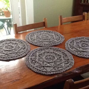 May include: Four grey crocheted placemats with a circular design on a wooden table.